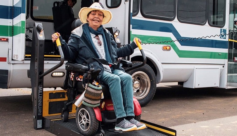 Woman using motorized wheelchair is lowered off a paratransit bus using a wheelchair lift.