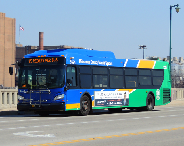 A Milwaukee County Transit System bus in operation, traveling westbound over the Wisconsin Avenue bridge with Miller Brewery in the background with a "15 riders per bus" message due to COVID.